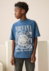 Sininen puuvillapaita, jossa on graafinen kuvio iloisella kasvoilla, teksti "NIRVANA" ja "COME AS YOU ARE" valkoisella. Lyhyet hihat ja rento istuvuus.