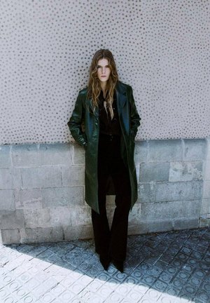 Green leather trench coat with structured lapels, paired with a black shirt and pants, set against a textured gray wall.
