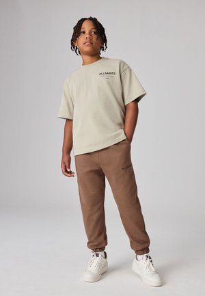 Ragazzo con i capelli intrecciati che indossa una t-shirt beige AllSaints, pantaloni da jogging marroni e sneakers bianche, in piedi con una mano in tasca.