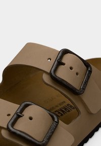 Κοντινό πλάνο λουριών μπεζ σανδαλιών με μαύρες ορθογώνιες μεταλλικές αγκράφες και ορατή την επωνυμία «BIRKENSTOCK» στην επένδυση του παπουτσιού.