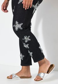 Jeans de mezclilla oscura con parches desgastados en forma de estrella, combinados con sandalias deslizantes plateadas que tienen una hebilla y una suela de corcho.