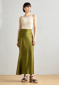 Beige top tank dipadukan dengan rok satin hijau zaitun panjang hingga lantai. Model mengenakan sandal hak kayu hitam dengan tali. Tekstur yang halus, desain minimalis.