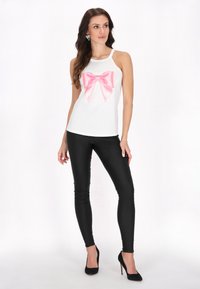 Fehér ujjatlan felső, nagy rózsaszín masni grafikával, fényes fekete leggingssel és fekete magassarkú cipővel párosítva. Sima szövettextúra.