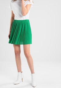 Groene geplooide rok gemaakt van lichtgewicht stof, gecombineerd met een wit t-shirt en witte enkellaarsjes met een spitse neus en hak.