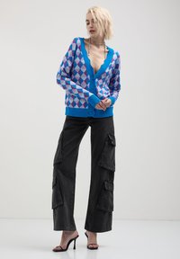 Cardigan blu con motivo a rombi rosa e viola, scollatura a V profonda, chiusura con bottoni, polsini e orlo a coste; abbinato a pantaloni cargo neri.