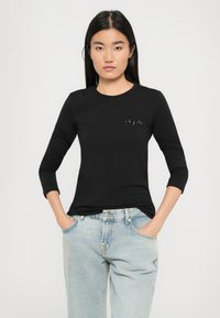 Schwarzes Langarmhemd mit rundem Halsausschnitt, versehen mit einem dezenten Logodetail auf der Brust, kombiniert mit hellblauen Denim-Jeans.