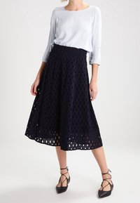 Gonna A-line blu navy con motivo a taglio, vita alta e tessuto fluido. Abbinata a una blusa azzurro chiaro e scarpe basse nere con lacci.