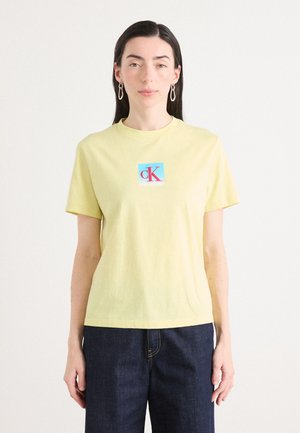 Lichtgele katoenen t-shirt met korte mouwen, met een vierkant logo met "cK" in roze en blauw in het midden. G gedragen met donkere jeans.