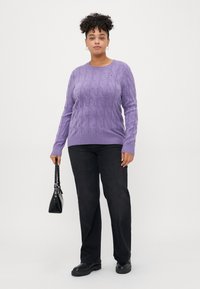 Lila Kabelstrickpullover mit langen Ärmeln, kombiniert mit schwarzen geraden Jeans. Das Model trägt eine kleine schwarze Handtasche.