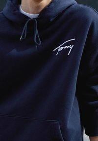 Marinblå hoodie gjord av mjukt tyg, med en framficka och en vit broderad "Tommy"-logotyp på bröstet, samt justerbara snören för huvan.