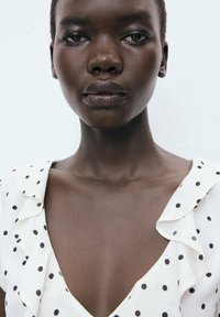 White dress with black polka dots, featuring ruffled edges. Model has short hair and smooth, dark skin, with a neutral background.