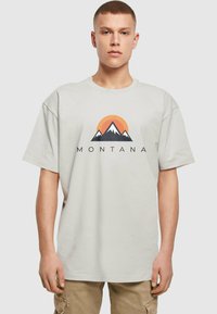 Camiseta de algodón gris claro con cuello redondo, que presenta un gráfico de montaña y el texto "MONTANA" en negro; corte relajado y mangas cortas.