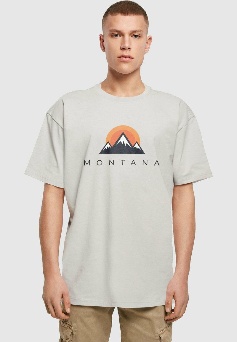 Camiseta de algodón gris claro con cuello redondo, que presenta un gráfico de montaña y el texto "MONTANA" en negro; corte relajado y mangas cortas.