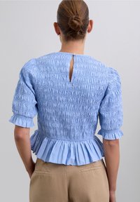 Woman with hair in a bun wearing a light blue smocked blouse with puff sleeves and ruffled hem, paired with beige pants, viewed from behind.