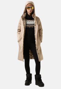 Persoon in een beige hoody lange jas, gestreept zwart sweater, zwarte leggings, zwarte laarzen, beige gebreide muts en zonnebril met gele tint.