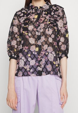 Elle porte un chemisier noir transparent avec un motif floral coloré et des détails froufroutants, associé à un pantalon violet clair avec des poches latérales.