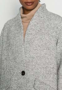 Szary płaszcz w jodełkę z kołnierzem szalowym, z czarnym guzikiem i otwartym przodem, noszony z jasnobeżowym golfem pod spodem.