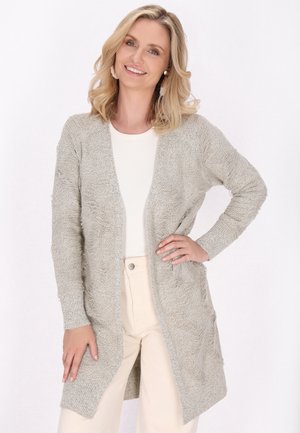 Femeie zâmbitoare cu păr blond poartă un cardigan texturat bej deschis peste un top alb și pantaloni crem, stând cu o mână pe șold.