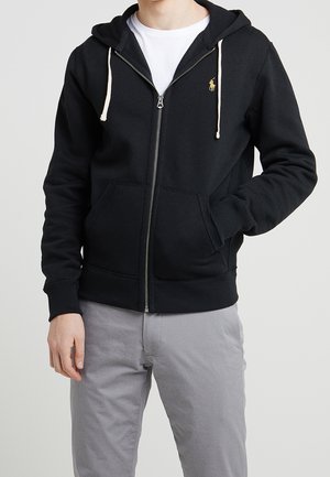 Homme portant un sweat à capuche noir avec fermeture éclair, cordons blancs et petit logo doré sur la poitrine, pantalon gris et t-shirt blanc en dessous.