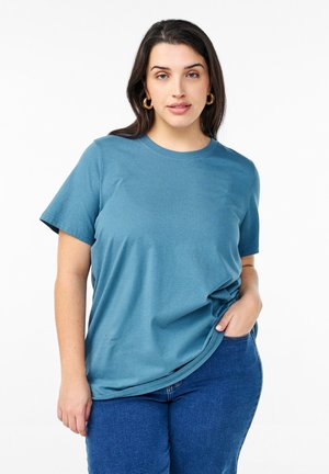 Teal cotton t-shirt with short sleeves and a round neckline. Slightly loose fit. Model wears it with blue jeans, hands in pockets.