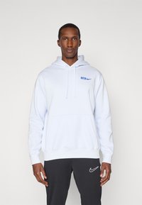 Svetlomodrá mikina s kapucňou a predným vreckom, na ktorej je nápis "INTER" a logo Nike v modrej farbe. Nosí sa s tmavošedými nohavicami, ktoré majú tiež logo Nike.