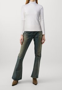 Weißes langärmliges Rollkragenoberteil kombiniert mit hochsitzenden, hellen bis dunklen Schlagjeans und braunen spitz zulaufenden Ankle Boots.