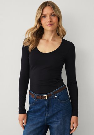 Kvinde med krøllet blondt hår iført en sort top med lange ærmer, stoppet ned i blå jeans med et brunt bælte, stående mod en ensfarvet baggrund.