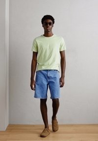 Lys grønn t-skjorte med korte ermer og blå denimshorts. Brune slip-on sko. Står mot en nøytral bakgrunn med tregulv.