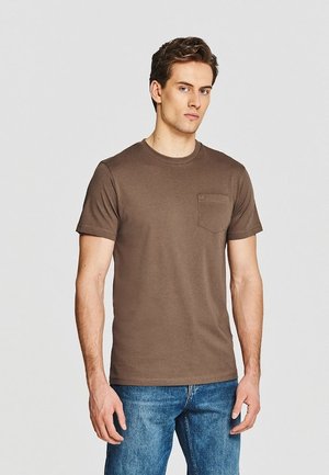 Młody mężczyzna w brązowej koszulce z krótkim rękawem i kieszenią oraz niebieskich dżinsach, stojący na tle jednolitego jasnego tła.