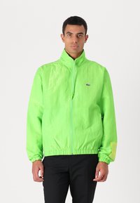 Casaco leve, verde néon, com fecho de zip, gola alta, punhos elásticos e bainha; apresenta um logótipo no lado esquerdo do peito e um detalhe amarelo na manga.