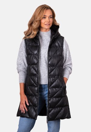 Woman wearing a black quilted sleeveless hooded vest over a gray sweater and blue jeans, standing with hands in pockets.