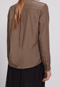 Camicia marrone a maniche lunghe con una texture liscia, orlo arrotondato e polsini abbottonati. Presenta un yoke sul retro e si drappeggia delicatamente sul corpo.