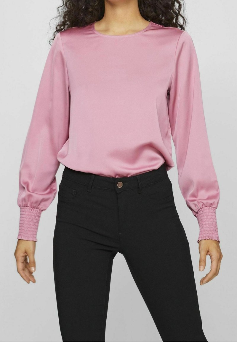 Blouse en satin rose avec manches longues bouffantes, dotées de poignets froncés. Associée à un pantalon noir ajusté. Texture lisse et encolure ronde simple.
