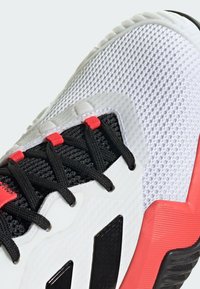 Weißer Sportschuh mit einem strukturierten Mesh-Obermaterial und schwarzen Schnürsenkeln; mit roten Akzenten und einem schwarzen Logo. Dicke Gummisohle für besseren Halt.