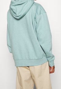 Felpa verde menta con una consistenza morbida, vestibilità oversize e cappuccio con lacci. Abbinata a pantaloni beige con polsini e orlo dettagliati.