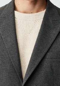 Blazer gris avec revers crantés, finition en laine texturée sur un pull en tricot crème. Poche visible sur le côté gauche du blazer.