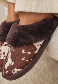 Kapcie w brązowo-kremowy wzór w krowie cętki, z miękkim, ciemnobrązowym futerkiem i teksturowanym wnętrzem. Wytrzymała podeszwa z obszytymi krawędziami.