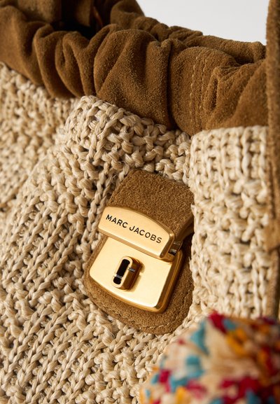 Κοντινό πλάνο υφαντής μπεζ τσάντας με καφέ σουέντ περιθώριο και χρυσό κούμπωμα χαραγμένο με το όνομα της μάρκας Marc Jacobs.