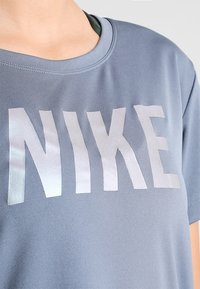 Blå atletisk t-shirt gjord av ett slätt tyg med en stor reflekterande silverfärgad "NIKE"-logotyp på framsidan, med rund halsringning.