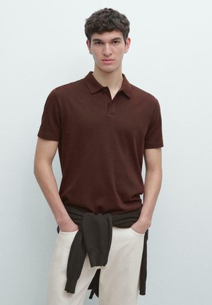 Mladý muž s kudrnatými vlasy, oblečený v krátkém hnědém polo tričku a bílých kalhotách, s tmavým svetrem uvázaným kolem pasu.