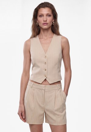 Vrouw draagt beige mouwloos vest met krijtstreep en knopen, en bijpassende hooggetailleerde shorts, staand met één hand in de zak tegen een witte achtergrond.