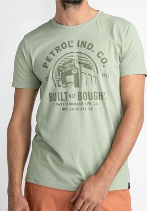 Hombre con camiseta verde claro con el logo de Petrol Ind. Co., una ilustración de garaje y el texto "Built Not Bought" en impresión marrón.