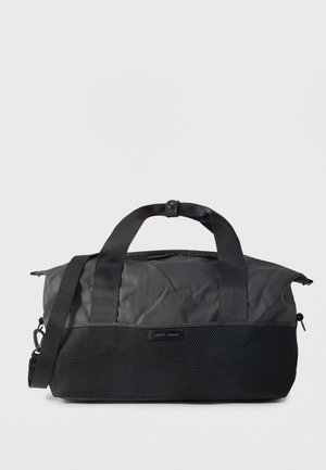 Sac de sport noir avec une poche avant en mesh, de courtes poignées et une bandoulière amovible, sur fond clair.