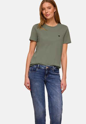 Młoda kobieta w oliwkowozielonym, krótkim rękawie z małym czarnym sercem jako emblematem i niebieskich wyblakłych dżinsach, stojąca w swobodnej pozie.