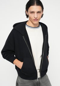 Sweat à capuche zippé noir avec une grande capuche, poches avant et poignets côtelés. Porté sur une chemise blanche avec un col rayé bleu.