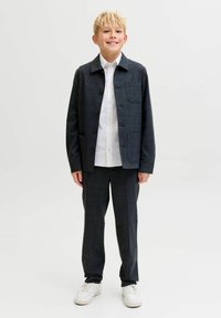 Vaikas dėvi tamsų languotą švarką su saga, po juo baltą marškinius ir derančias kelnes, derinamas su baltomis sportinėmis batelėmis.