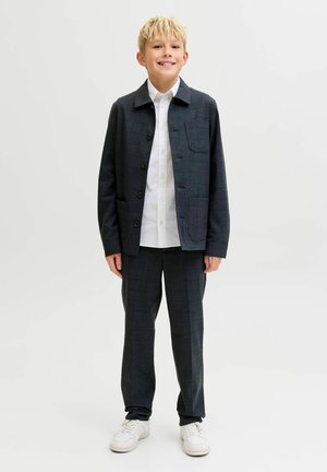 Barn kledd i en mørk rutete dressjakke med knapper, en hvit skjorte under og matchende bukser, kombinert med hvite joggesko.