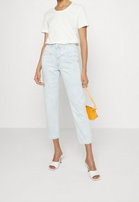 Person som bär en vit t-shirt, ljusblå högmidjade jeans, vita klackslippers och håller en liten gul handväska i remmen.