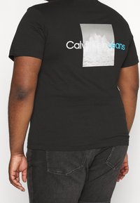 T-shirt nera in cotone con una grafica prominente di onde e il testo "Calvin Klein Jeans" in bianco e blu. Maniche corte e collo rotondo.