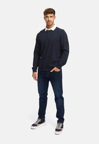 Granatowa koszula polo z długim rękawem i białym kołnierzykiem, zestawiona z ciemnoniebieskimi dżinsami oraz czarnymi trampkami z szarymi akcentami. Prosty, casualowy design.